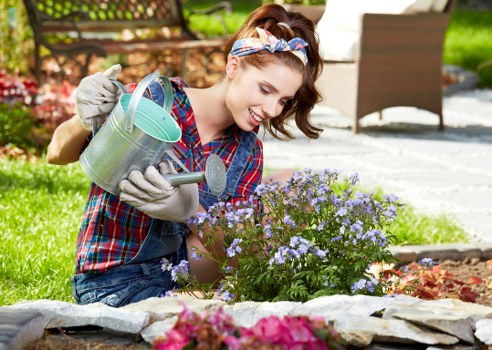 A woman with light skin, brown hair tied back with a floral headband, wearing a colorful plaid shirt and grey gardening gloves, is kneeling in a landscaped garden in Hoxton. She is watering a thriving shrub with small, purple flowers using a silver metal watering can. The garden features a neatly edged flower bed with a variety of plants, including pink and red flowers, laid out on a stone or concrete border. In the background, there is a well-maintained lawn with lush green grass, a wooden patio table and chairs, and some additional garden beds or plantings, indicating a thoughtfully designed outdoor space. The weather appears bright and sunny, with natural daylight illuminating the scene, highlighting the vibrant greenery and flowering plants. This detailed depiction aligns with gardening services such as maintenance, planting, and garden clearance offered by Gardening Services Hoxton, reflecting a well-kept suburban garden environment in the local area.