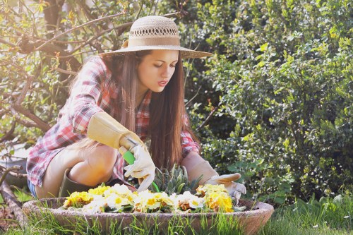 A young woman wearing a wide-brimmed straw hat, plaid shirt, and gardening gloves is kneeling in a lush garden with vibrant green foliage and sunlight filtering through trees. She is carefully tending to colorful flower beds, which include yellow, white, and purple blooms, arranged in a curved planting bed bordered by a low stone or brick edge. The grass surrounding the flower beds is well-maintained and dense, with a mix of natural textures. In the background, there are tall shrubs and trees providing shade and creating a natural outdoor environment typical of a residential backyard garden in Hoxton. The scene suggests active outdoor gardening, aligning with professional gardening services such as maintenance, planting, and landscape care provided by Gardening Services Hoxton. The lighting indicates a clear, mild day, ideal for garden activities.