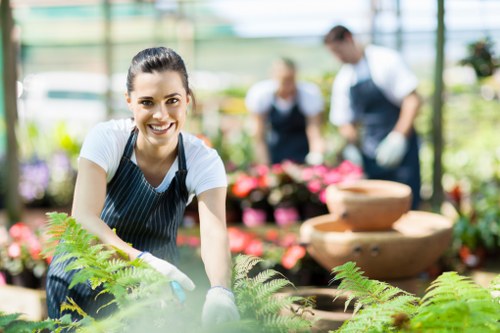 A young woman wearing a striped apron and white gloves is working in a greenhouse or garden centre, surrounded by various potted plants and flowers. She is leaning forward slightly with a smile, tending to a lush, green fern plant in the foreground. Behind her, there are blurred figures of a man and a woman, both dressed in gardening attire, attending to different plants amidst a vibrant display of colourful flowers and foliage. The environment is bright, filled with natural light filtering through glass panels, suggesting a warm, sunny day suitable for outdoor or garden maintenance. The scene emphasizes a well-organized, thriving garden environment, highlighting features such as healthy greenery, neatly arranged plants, and decorative garden features, consistent with a professional gardening service setting in Hoxton or nearby areas, supporting the availability of comprehensive maintenance, clearance, and landscaping services by Gardening Services Hoxton.
