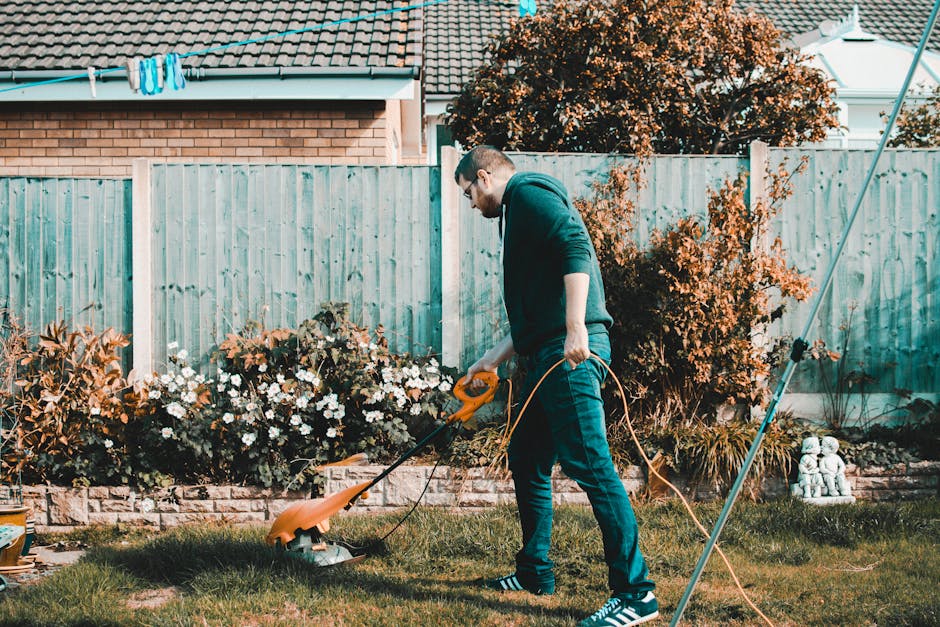A gardener wearing a dark long-sleeve shirt and a grey cap is trimming a dense, well-maintained hedge with long-handled shears. The hedge consists of small, glossy green leaves and is situated along a