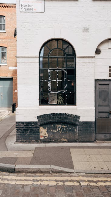 The image shows the exterior of a white brick building with an arched window, which features black framing and multiple panes, revealing a potted plant inside. Below the window is a weathered black br
